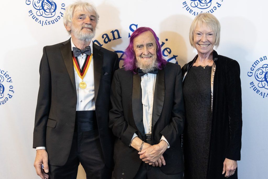 Three people in formal black attire standing on a backdrop at an event, smiling for a photo