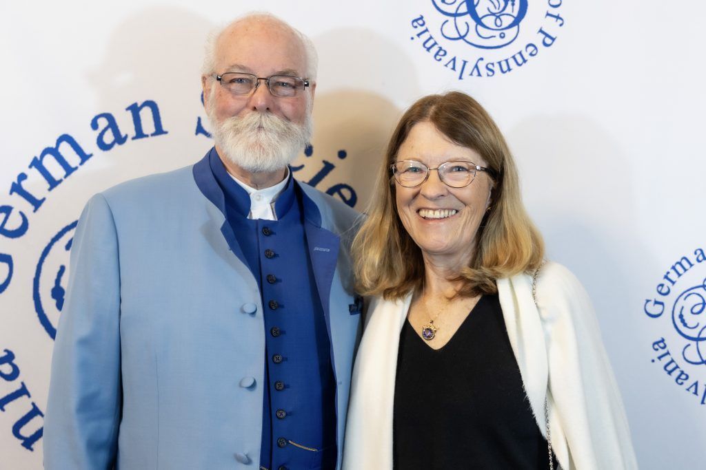 Two people smiling at a German-themed event backdrop, one in a blue vest and one in a white cardigan.