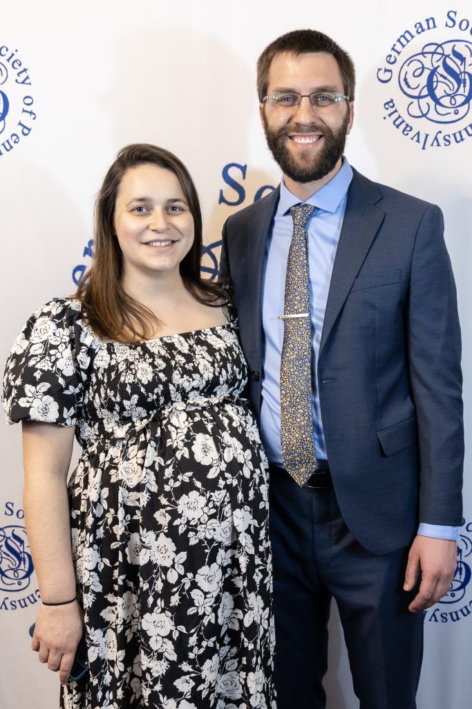 Two smiling people posing in front of a branded backdrop, wearing formal attire