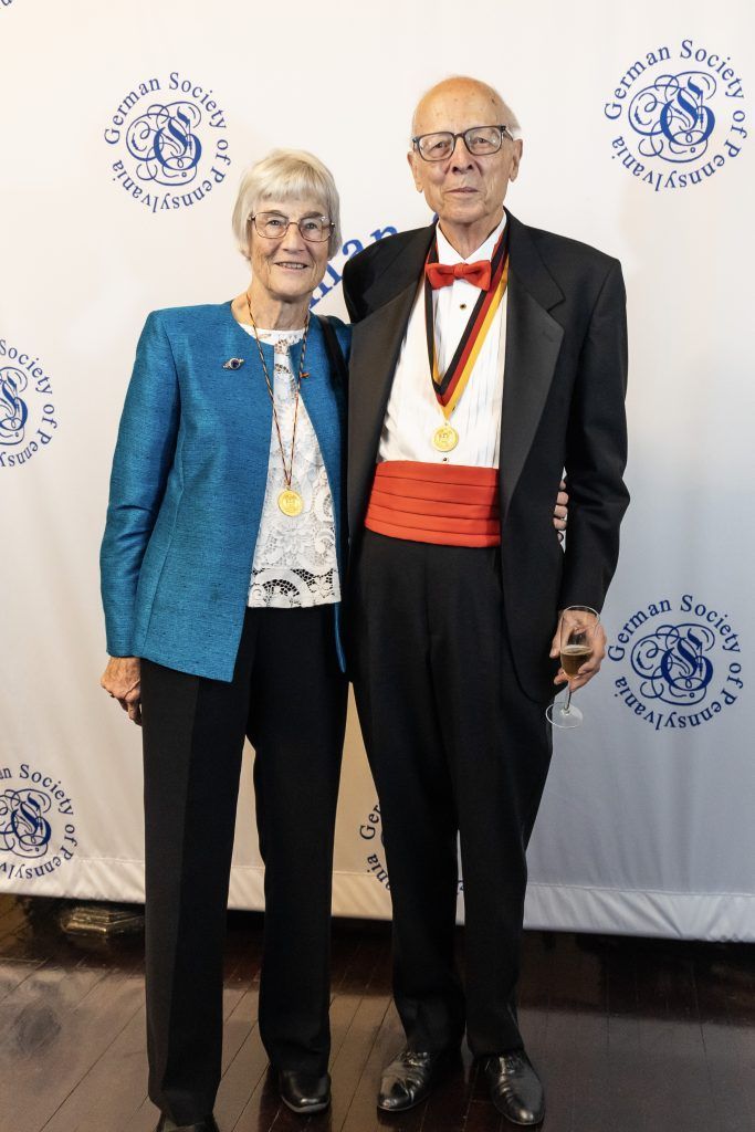 Two people posing in formal attire at a ceremony backdrop, one wearing a medal and red sash.