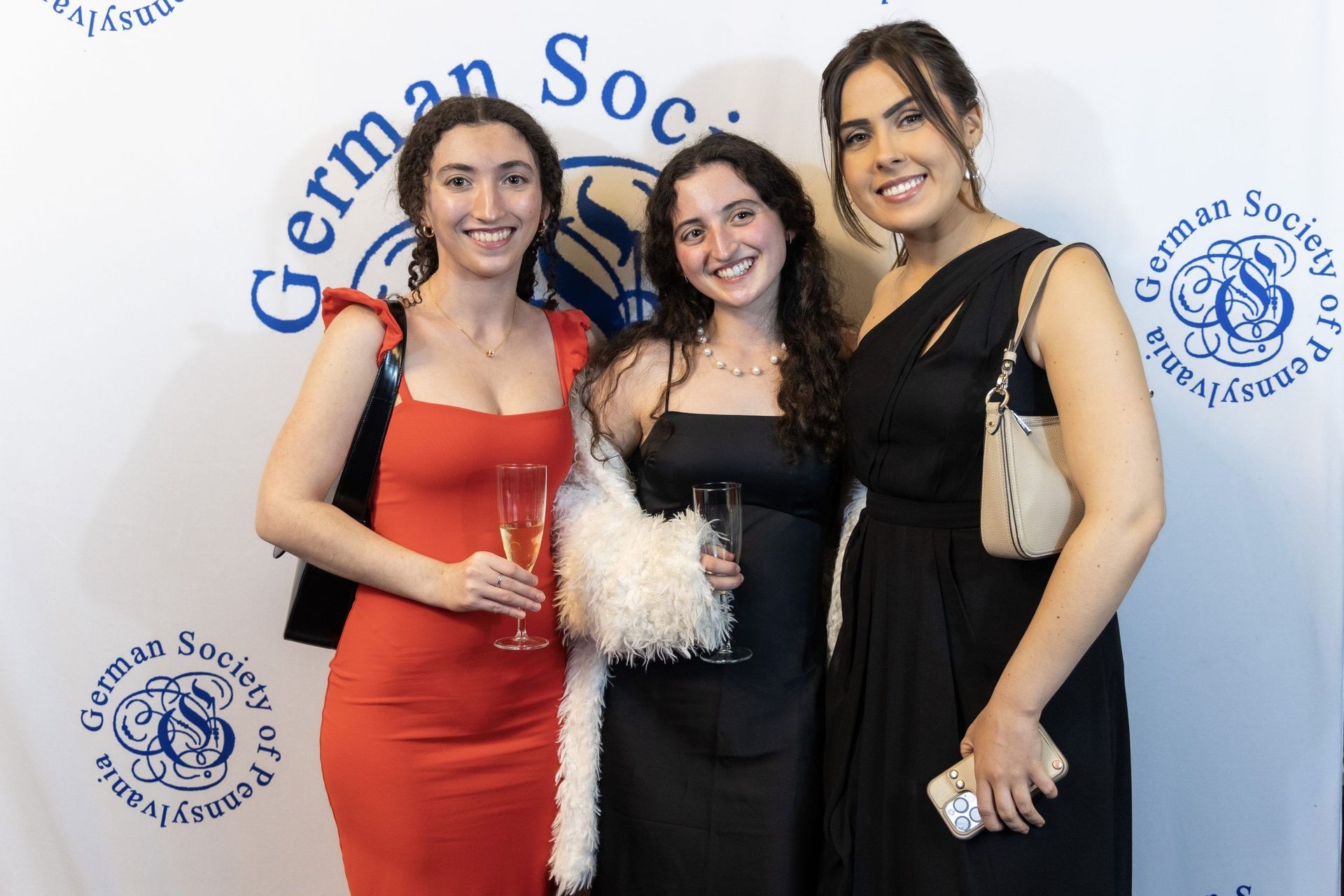 Three people posing in formal dresses at a German Society event backdrop
