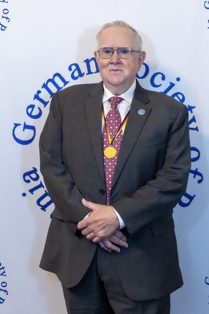 Man in a dark suit stands before a German Society backdrop, hands clasped, wearing a medal and patterned tie