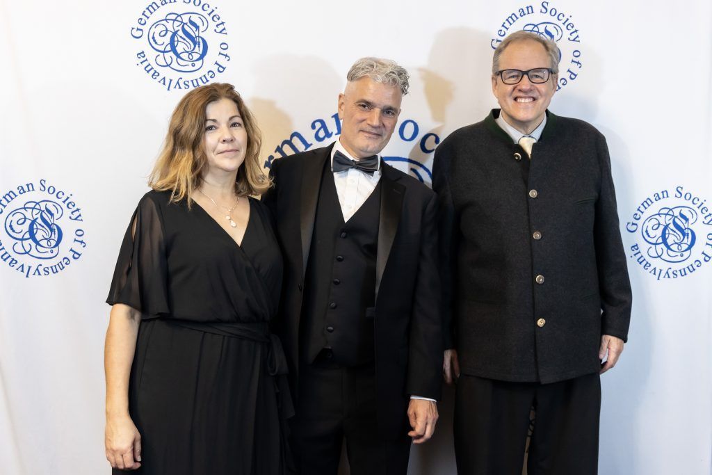 Three people posing in formal black attire at a branded event backdrop.