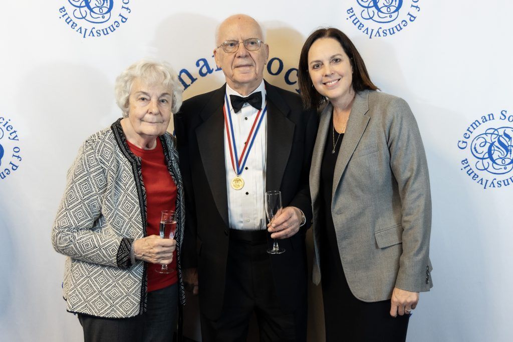 Three people posing at a formal event, with a medal worn by the man in the center.