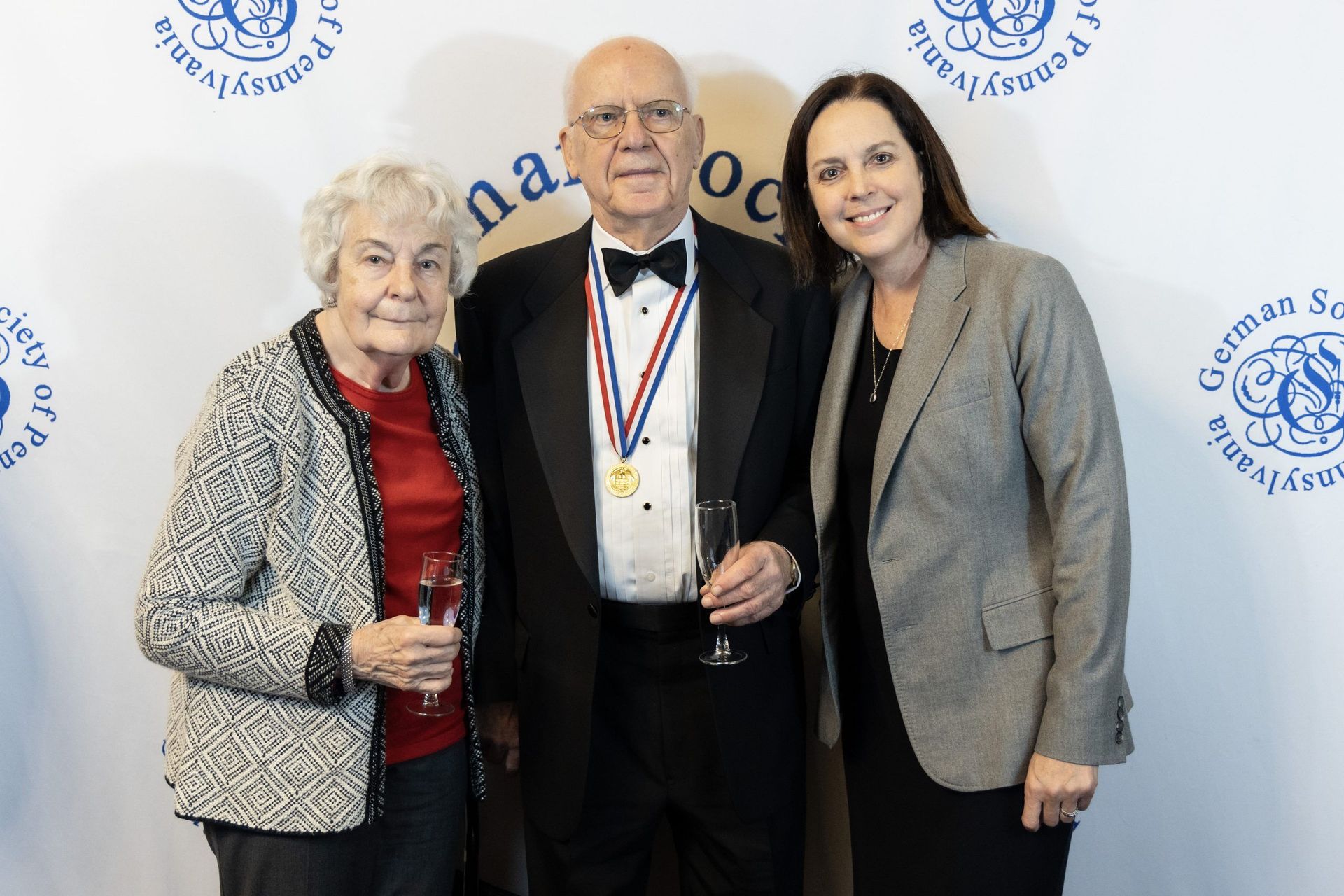 Three people posing in front of a backdrop, two women flanking a man wearing a medal.