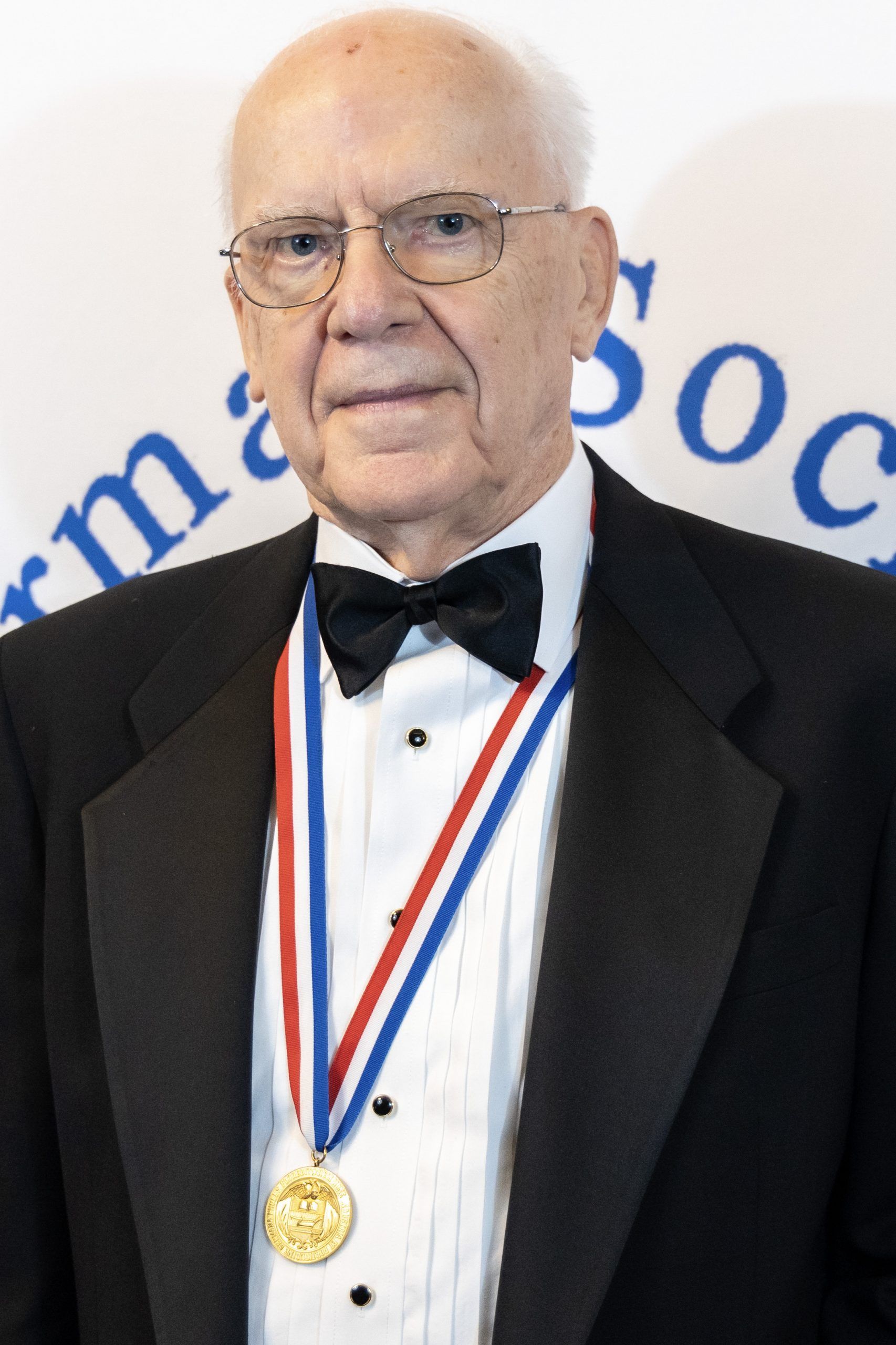 Man in tuxedo with medals, standing before a white backdrop with blue text
