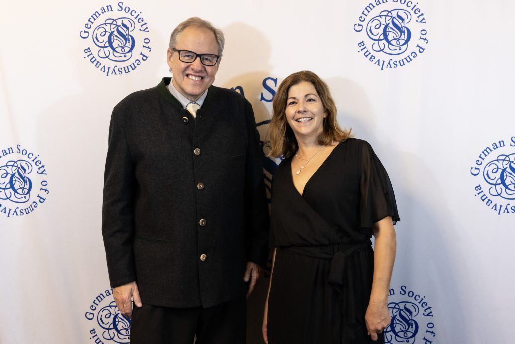 Two people smiling at a step-and-repeat event backdrop, dressed in dark attire