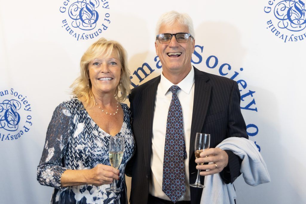 Two smiling guests at a formal event, standing before a branded backdrop with drinks in hand.