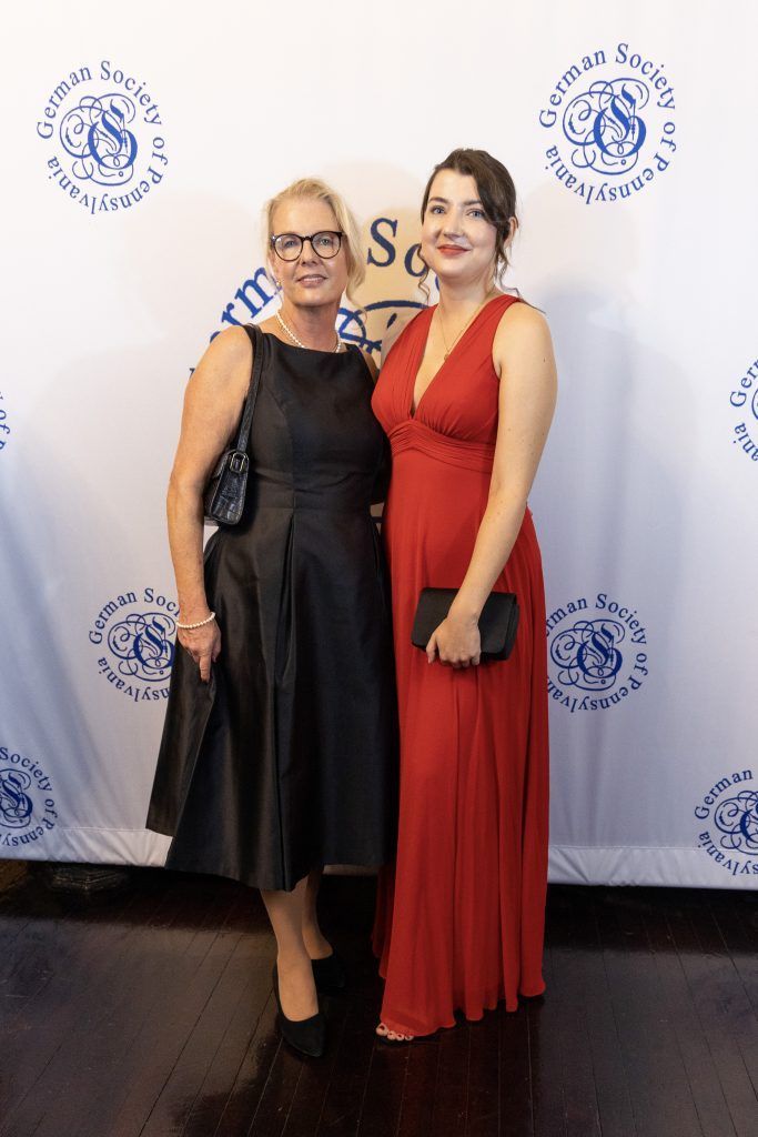 Two women posing on a step-and-repeat backdrop, one in a black dress and one in a red gown.
