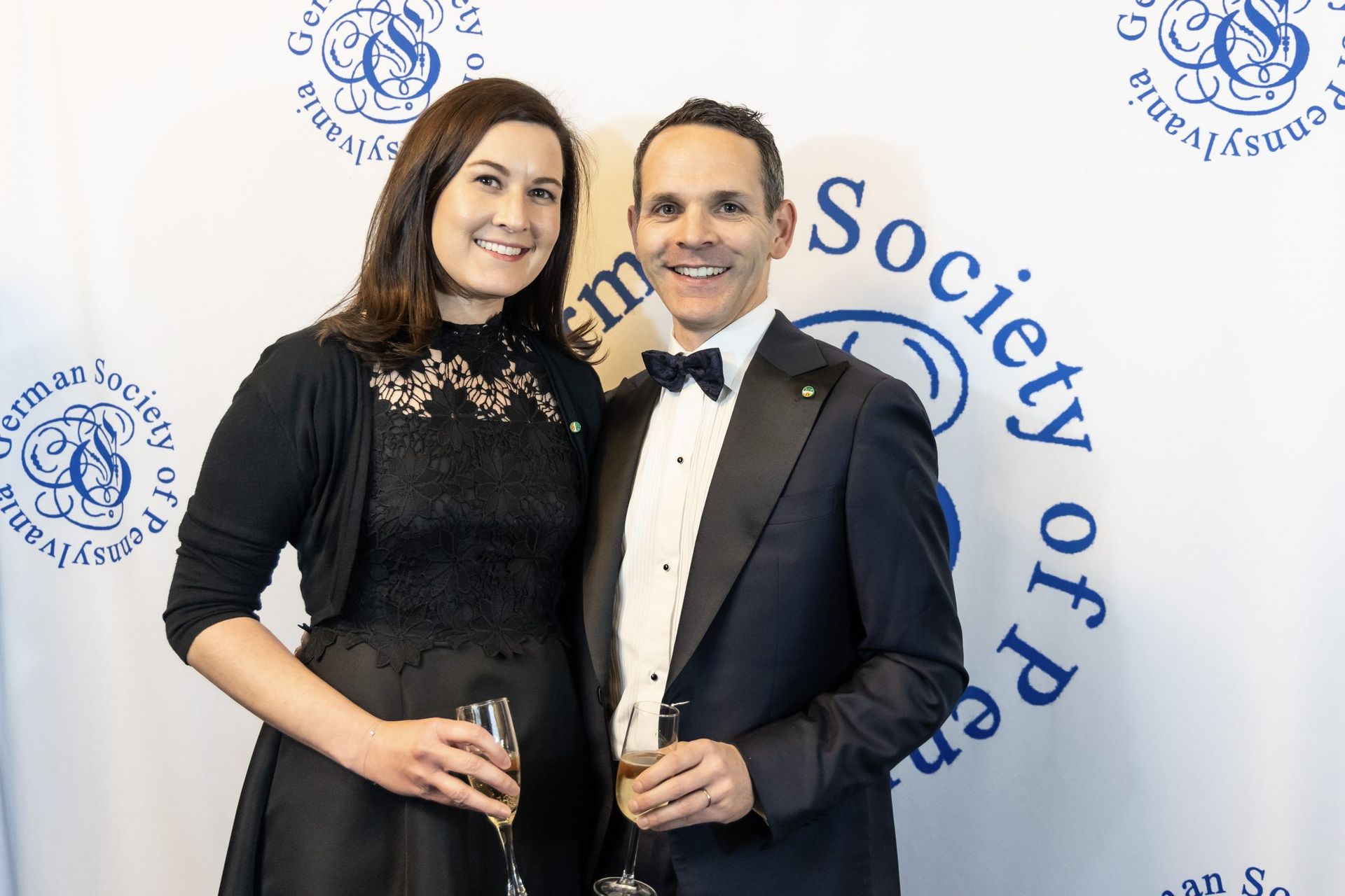 Two people posing at a Society of Professionals event, smiling and holding drinks before a logo backdrop