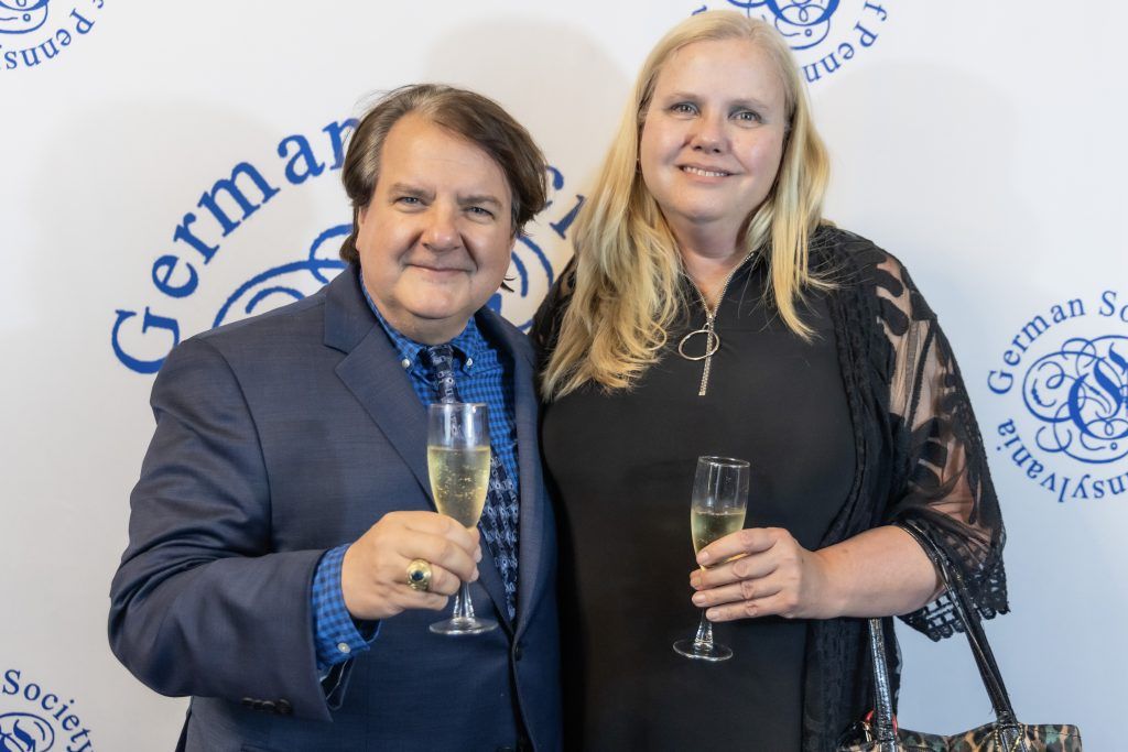 Two people smiling and holding champagne glasses at a German-themed event backdrop.