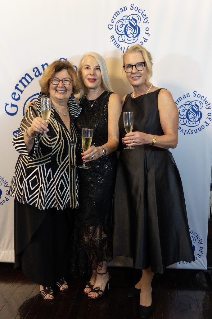 Three women in formal black outfits posing with drinks at a German-themed event backdrop