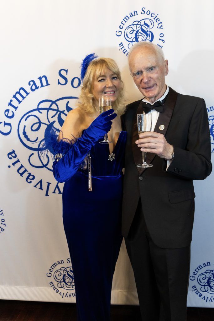 Two people posing at a formal event, one in a blue dress and one in a tuxedo, in front of a German Society backdrop