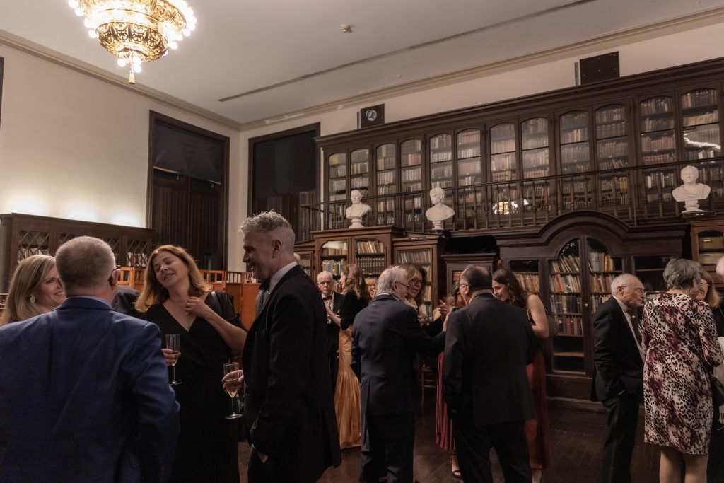 Guests mingling in a warmly lit elegant room with chandeliers and a large glass-front cabinet.