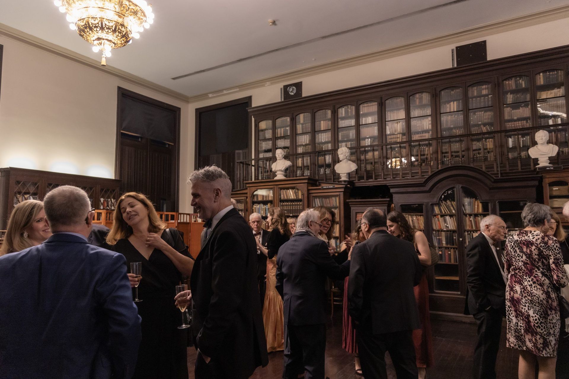 People mingling in a formal room with dark wood cabinets and a chandelier.