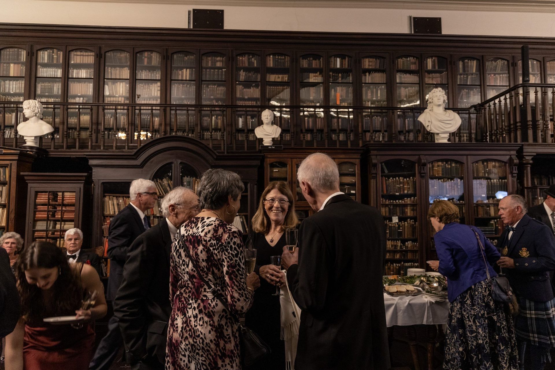 People mingling in a dim library, with tall bookshelves and a buffet table in the background.