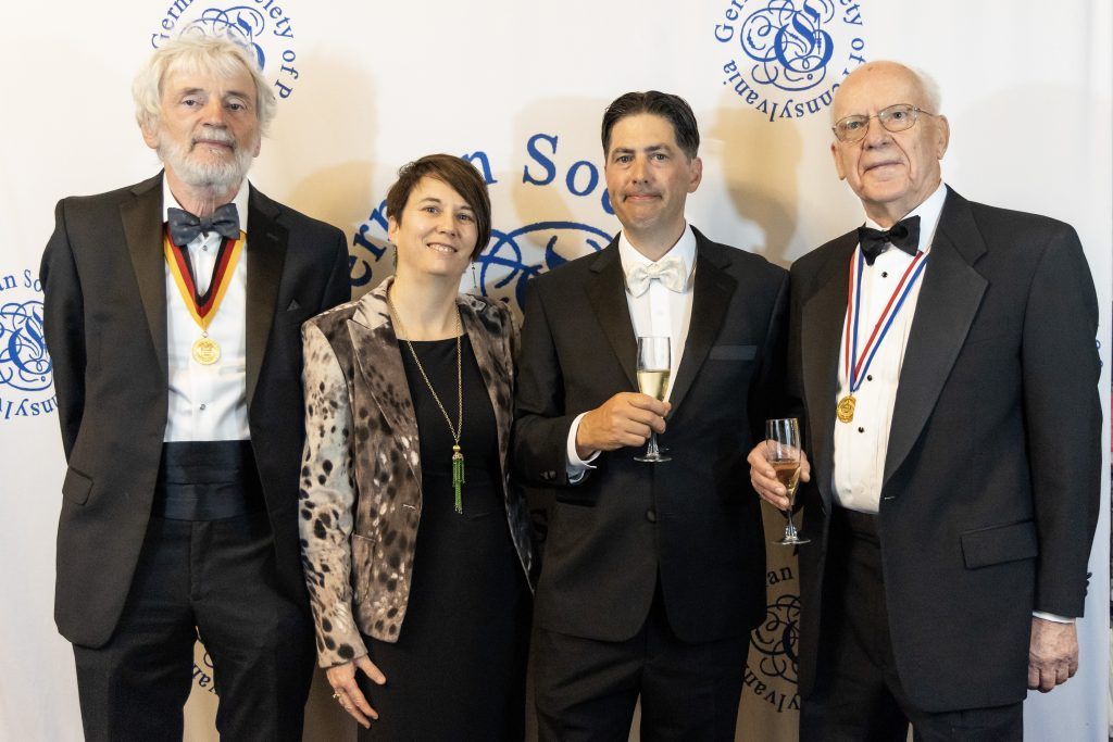 Four people in formal wear holding drinks, posing at a banquet or award event backdrop