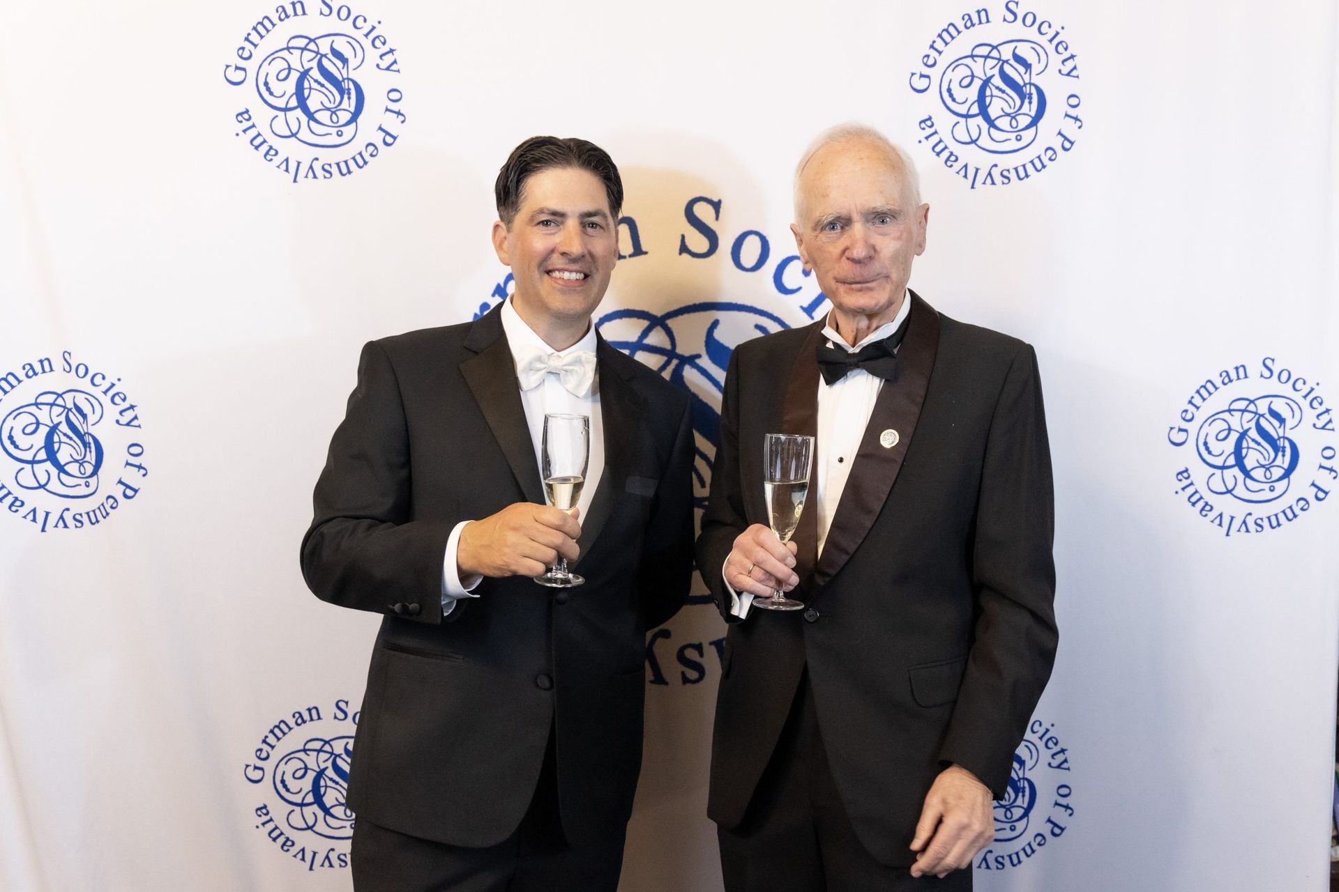 Two men in tuxedos holding awards at a formal event backdrop