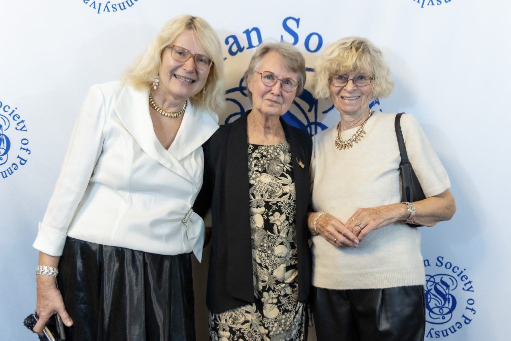 Three smiling people pose together at an event backdrop, one holding a black clutch.