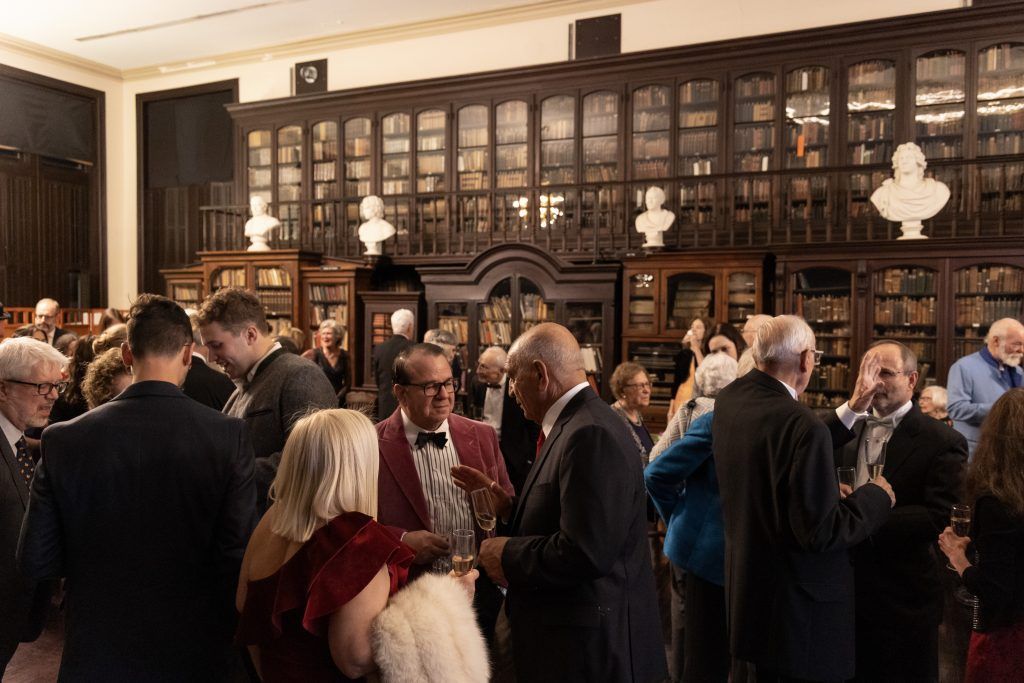 People mingling in a formal room with dark wood bookshelves and cabinets, holding drinks and chatting