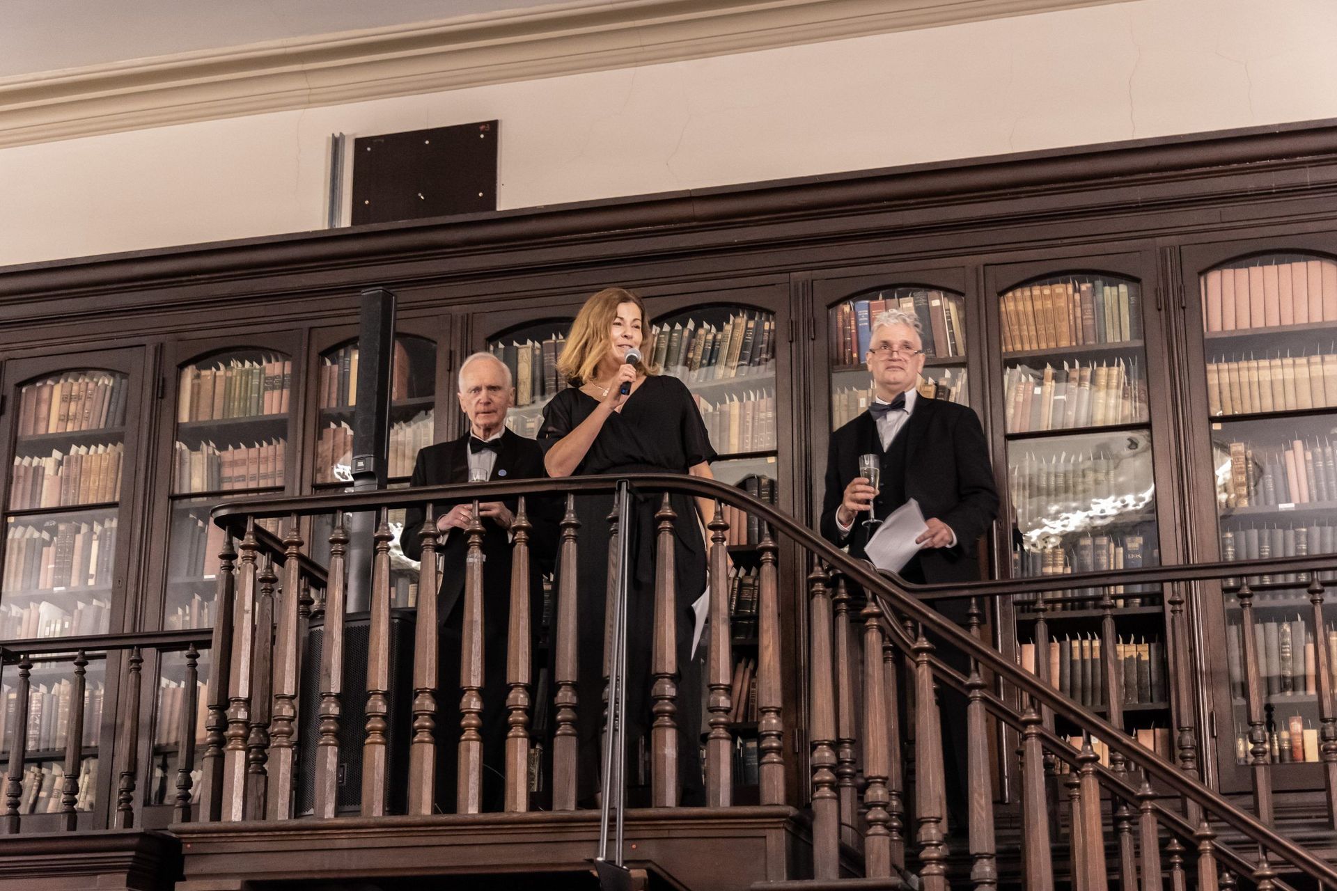 Three people stand on an ornate wooden balcony in a library lined with books.