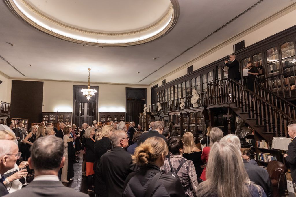 Crowded formal gathering in an ornate room with a balcony and chandelier, attendees facing the front.