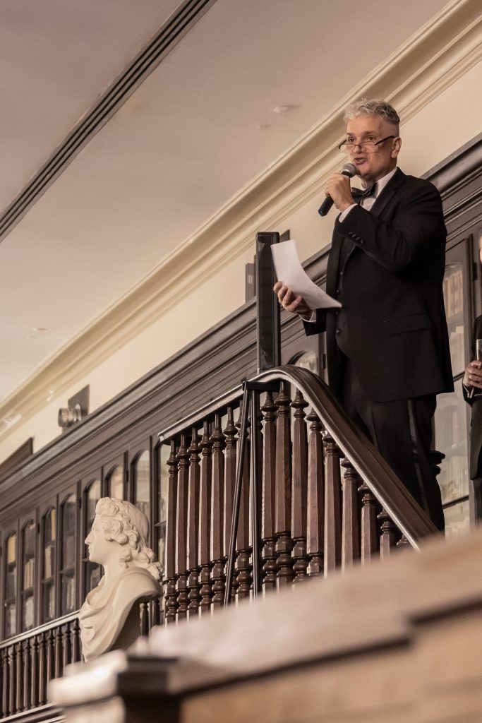 Man in dark suit speaking into a microphone beside a staircase and carved railing in a grand interior