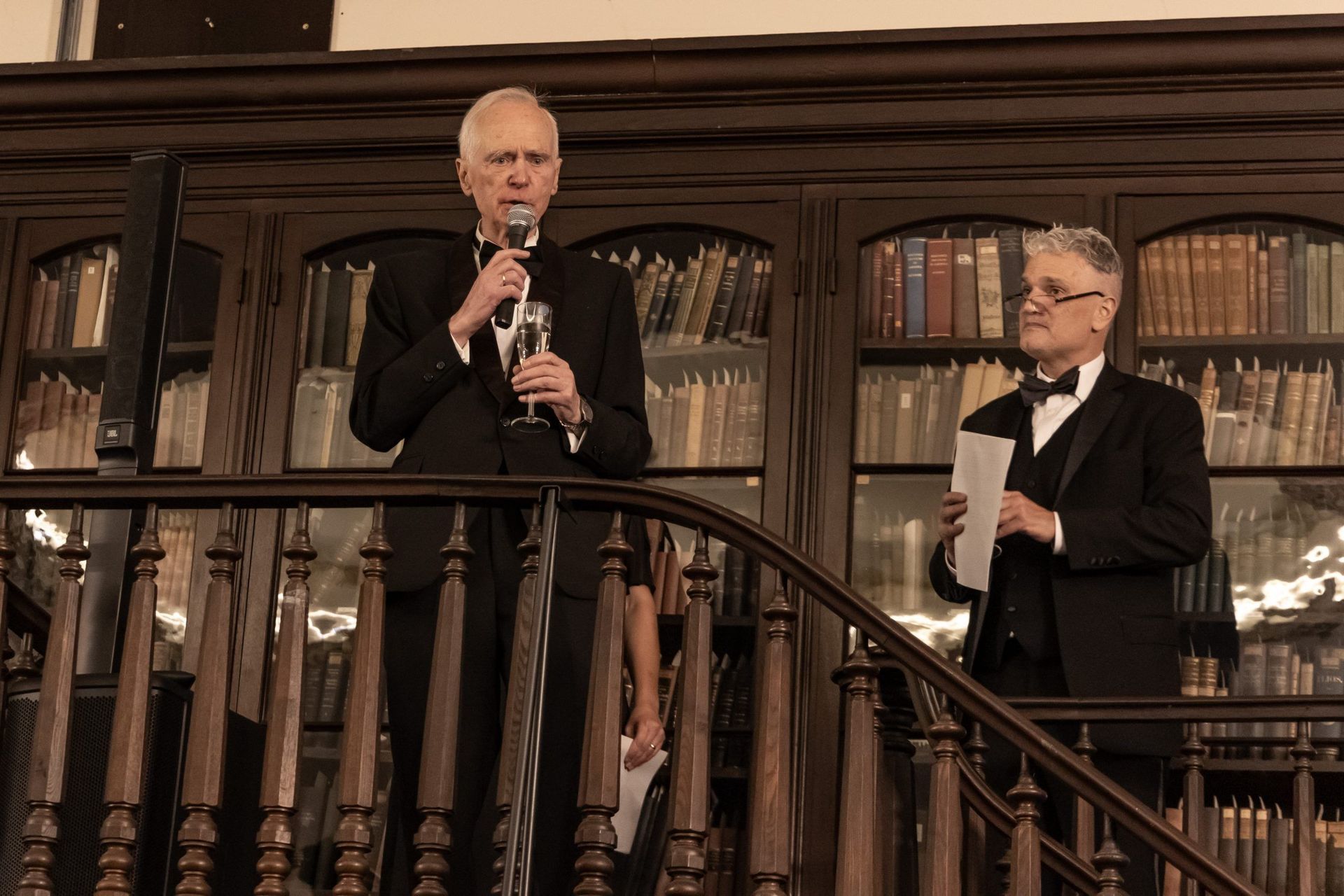 Two men in tuxedos stand on a balcony in a library, one speaking into a microphone