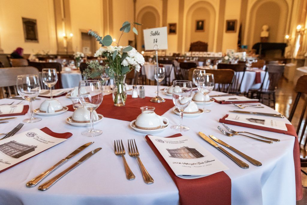 Formal banquet table set with white linens, red runner, glassware, silverware, and white floral centerpieces in a hall