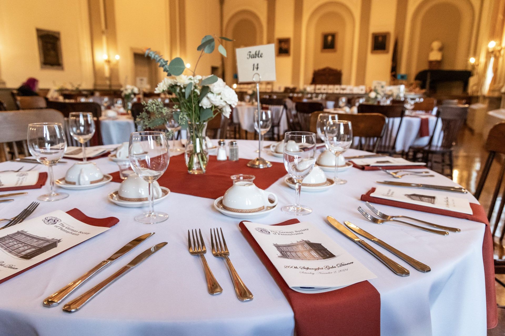 Elegant banquet table set with white linens, gold cutlery, red runners, and floral centerpieces in a grand hall
