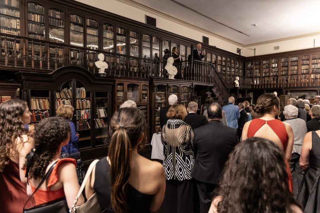 Crowded library reading room with people viewing tall wooden bookshelves and glass-front cabinets