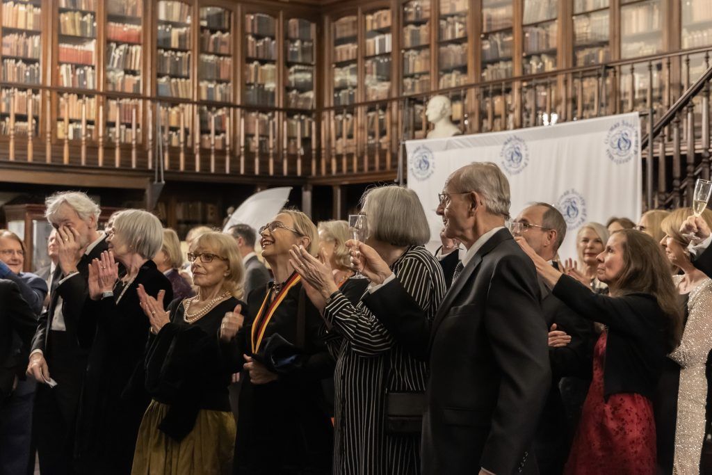 Audience applauding in a grand library hall with tall bookshelves and a white banner backdrop