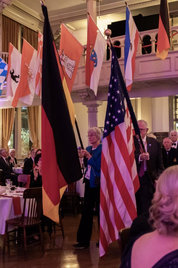 People carrying U.S. and German flags in a formal indoor ceremony with seated guests