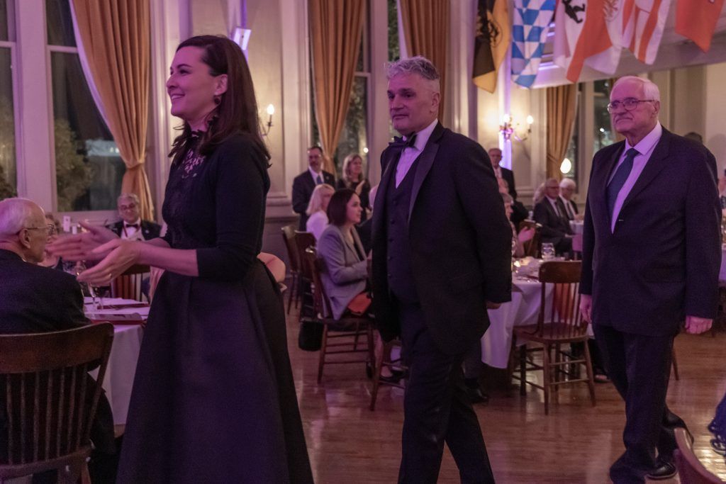 People in formal attire walking through a warmly lit banquet hall with flags hanging overhead
