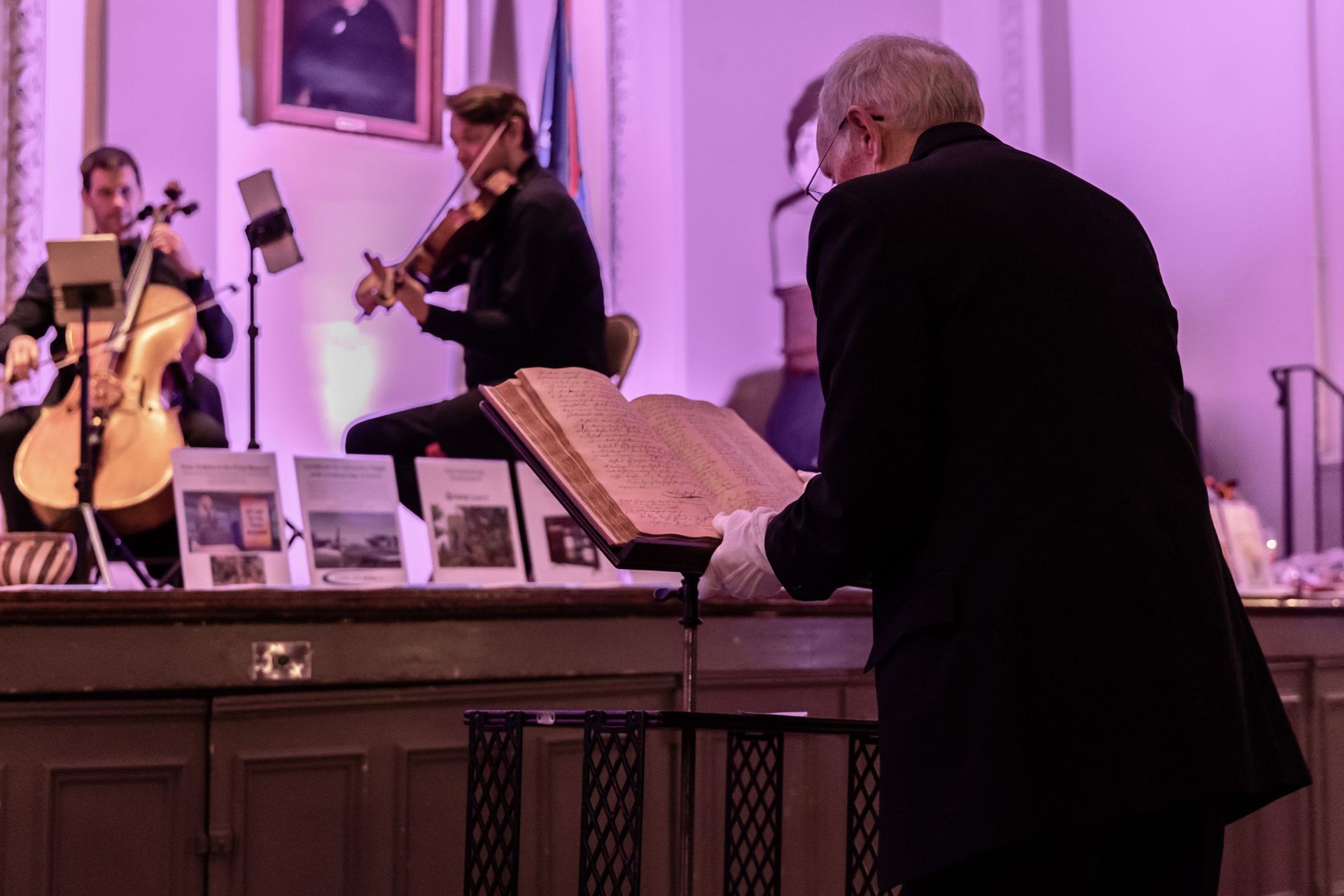 Musicians performing indoors while a conductor reads sheet music at a piano under pink lighting