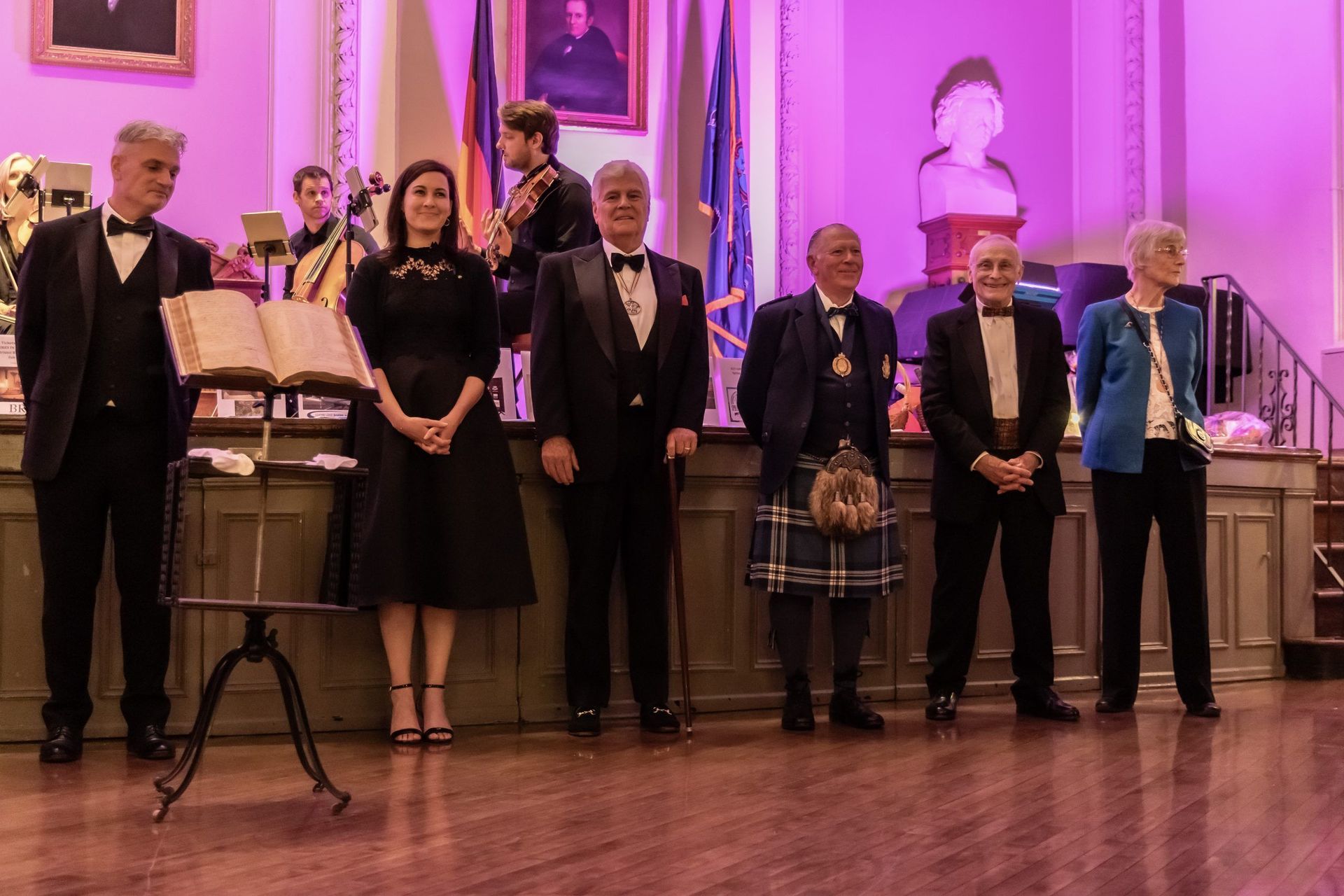 Six people standing on a stage at an awards ceremony under pink lighting