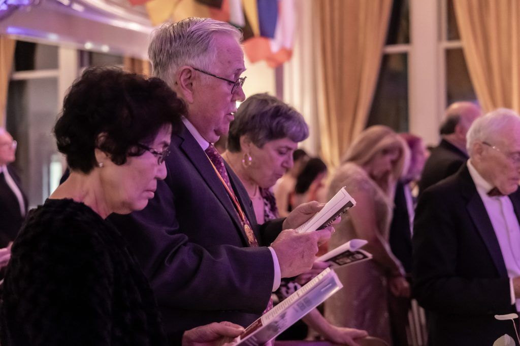 Audience members in formal attire reading programs at a softly lit event