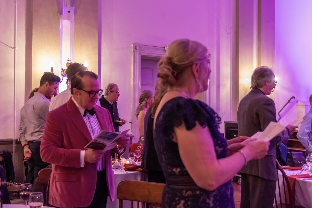 Guests holding papers at a softly lit indoor event with purple lighting and a table in the background