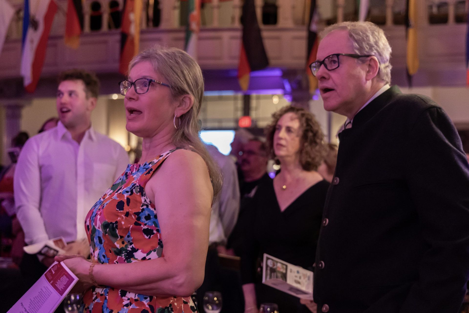 People in formal attire watching an event, with one woman in a colorful dress in the foreground.
