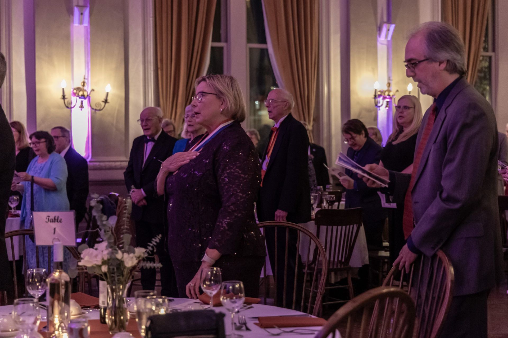 Guests stand at a formal banquet in a candlelit ballroom with gold drapes and purple lighting.