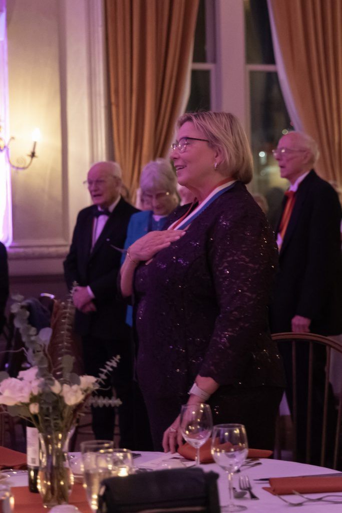 Guests standing and singing at a formal dinner with gold curtains and candlelit tables.