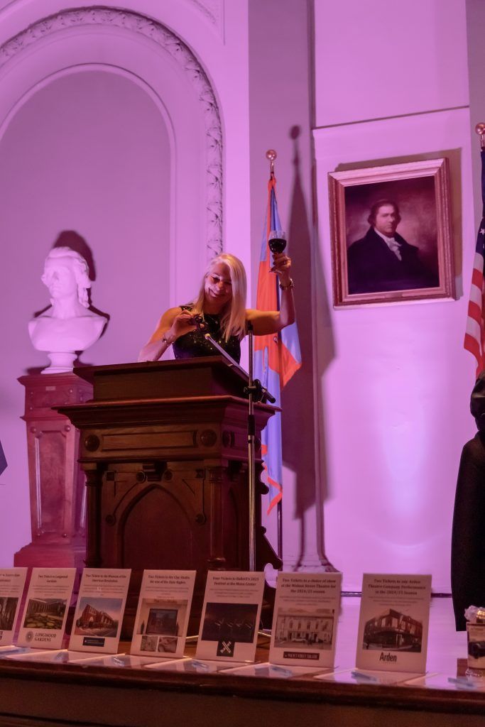 Person speaking at a podium in a museum-like room with portraits, busts, and informational placards.