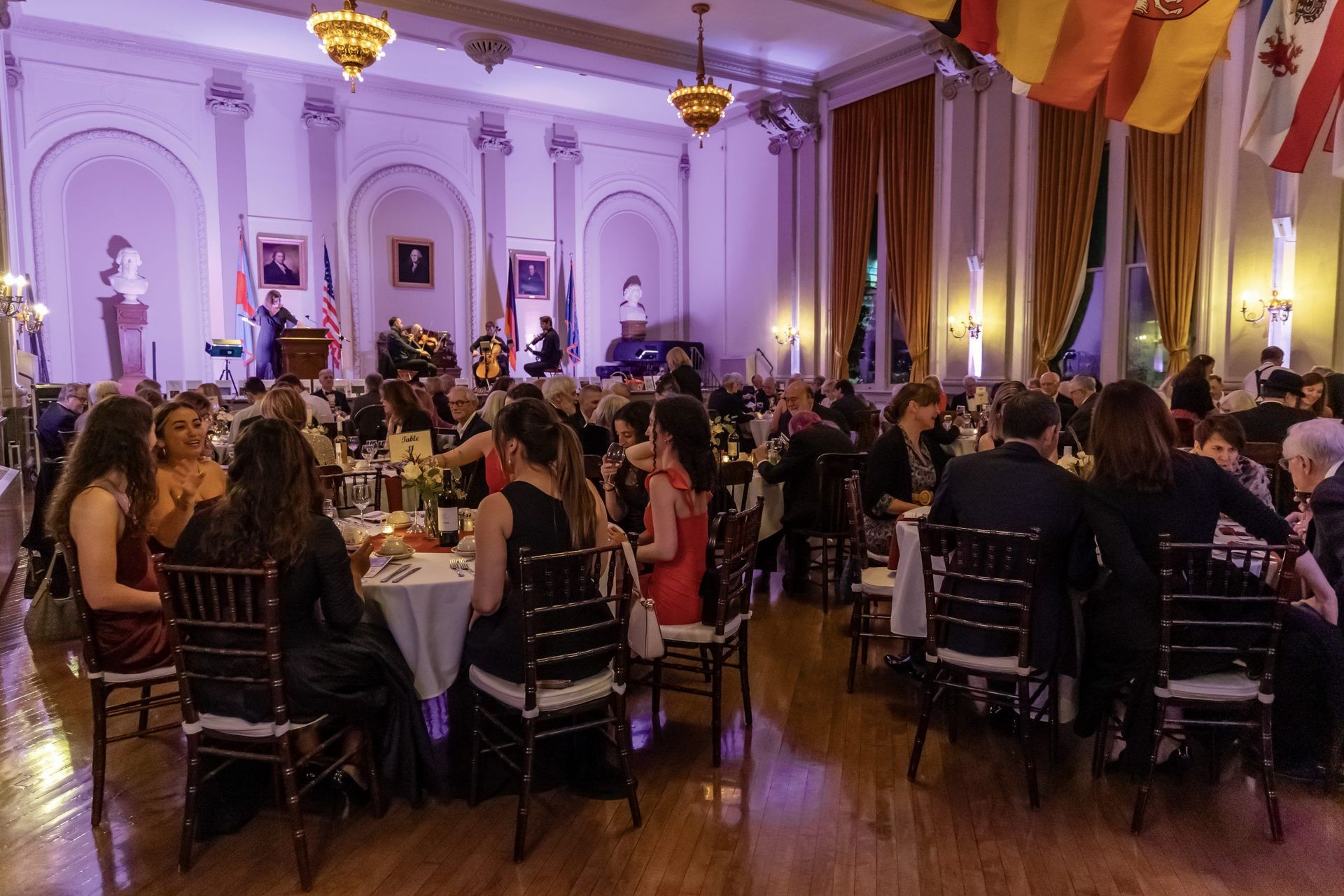 Guests seated at banquet tables in an elegant purple-lit ballroom with a stage and speaker at the front