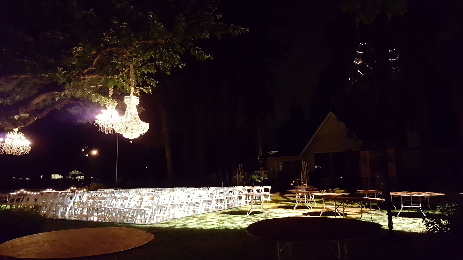 Outdoor evening event with chandeliers hanging from trees, chairs arranged in rows, and tables in the background.