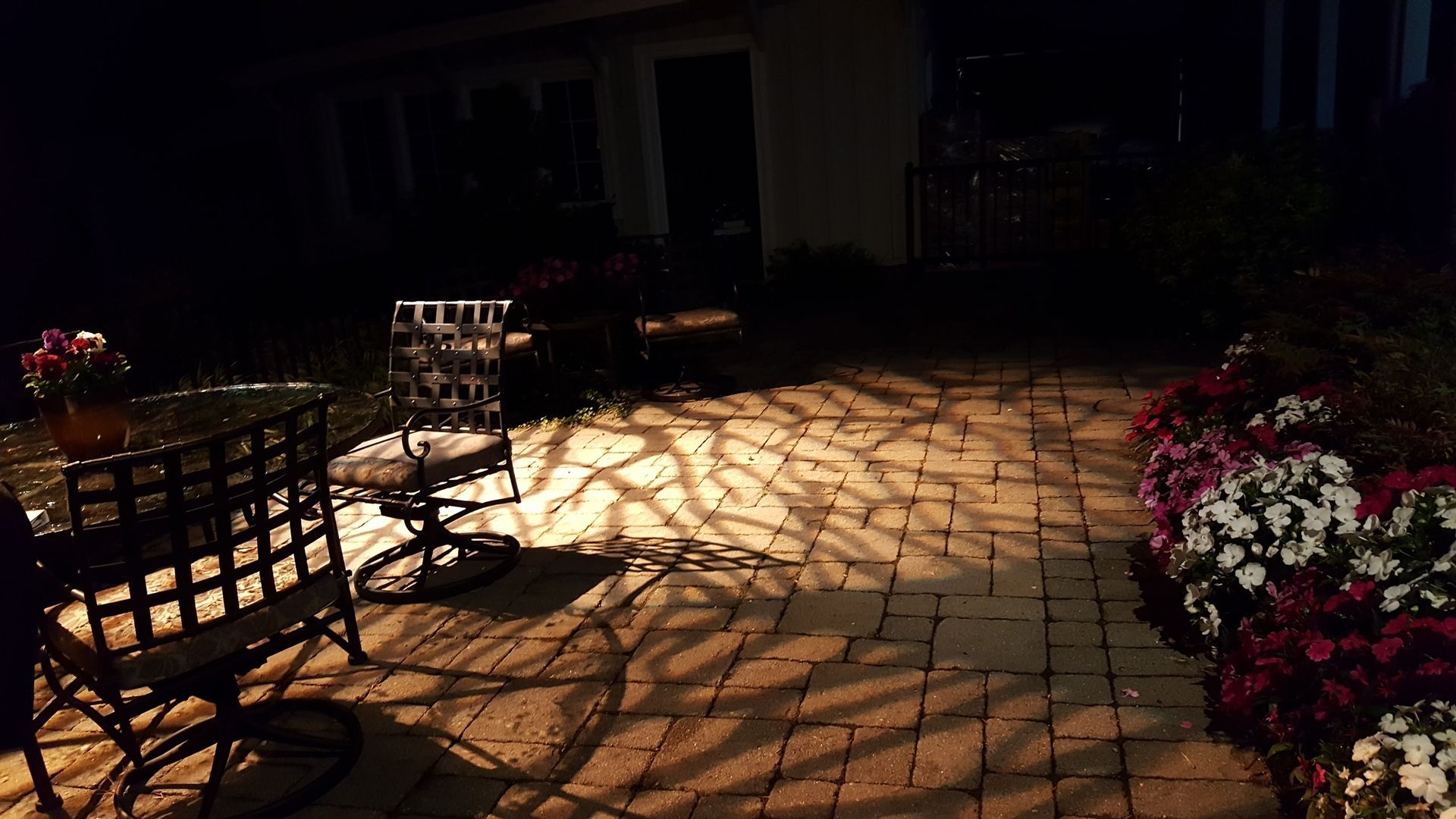 A patio at night with a table, chairs, and flowers. Light creates shadows on the brick ground.