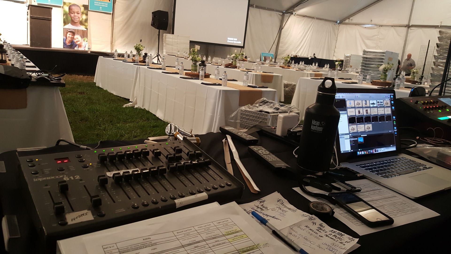 Sound mixing console and laptop set up for an event in a tent, with tables and a screen in the background.