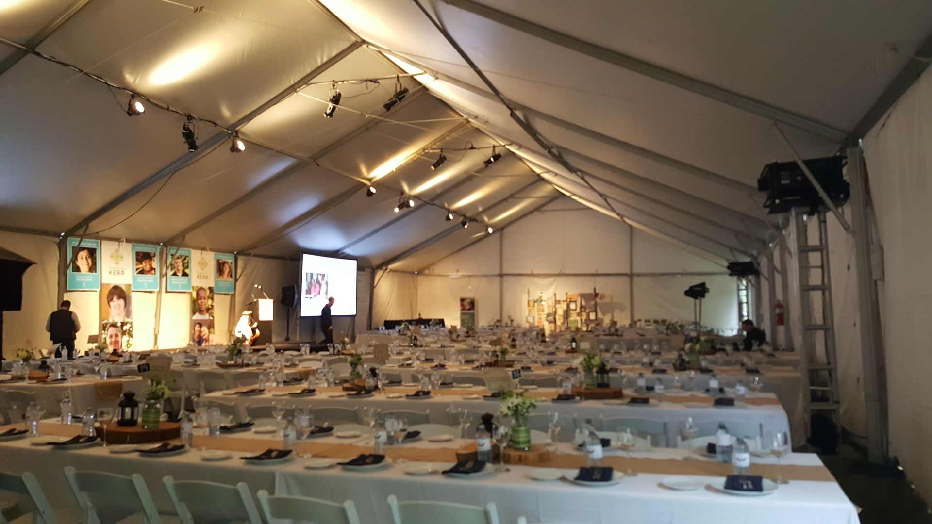 Inside a large white tent, tables set for a banquet. People mill about near a screen and artwork.