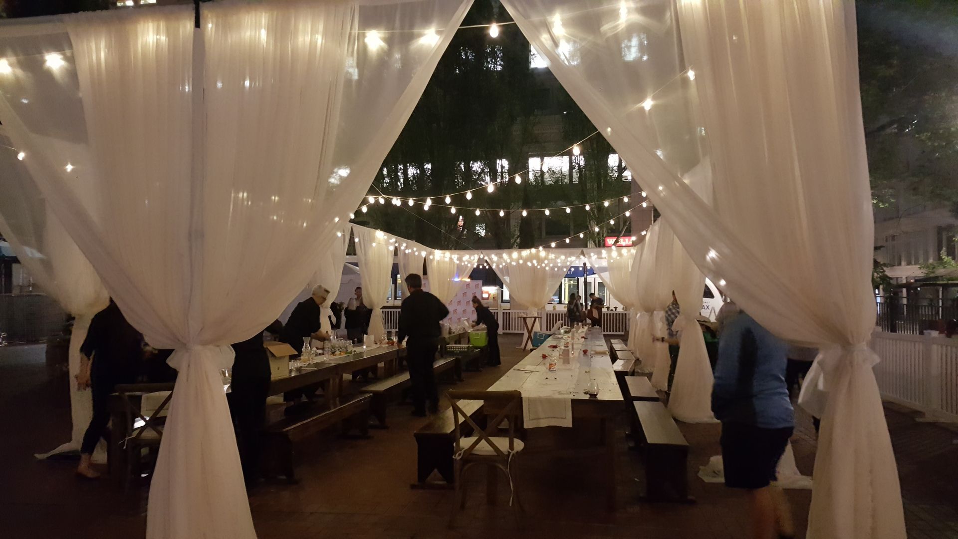Outdoor dining area with white draped fabric, string lights, and long wooden tables with people.