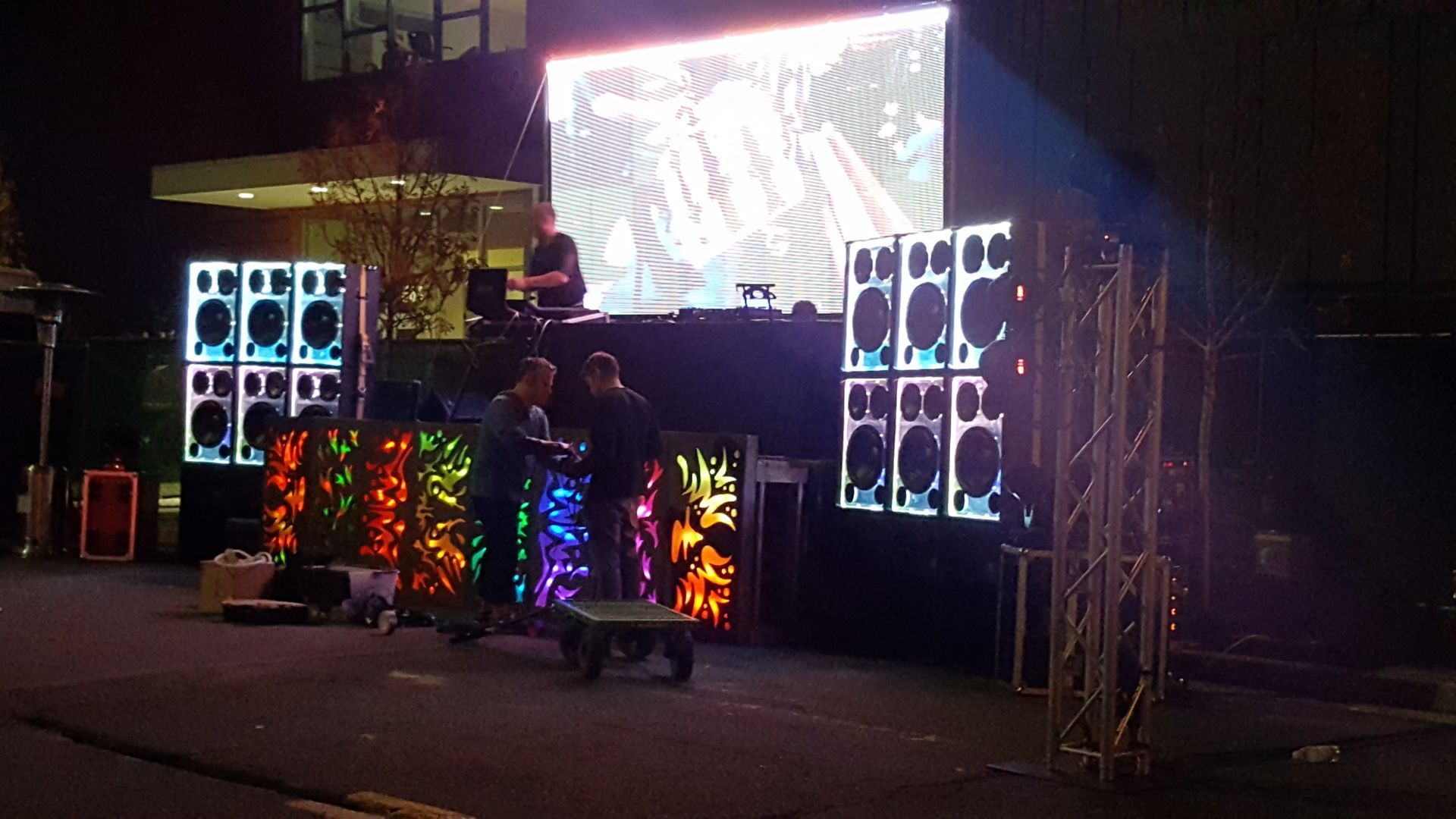 DJ setup with large screen and speakers on a street at night, with crew setting up equipment.