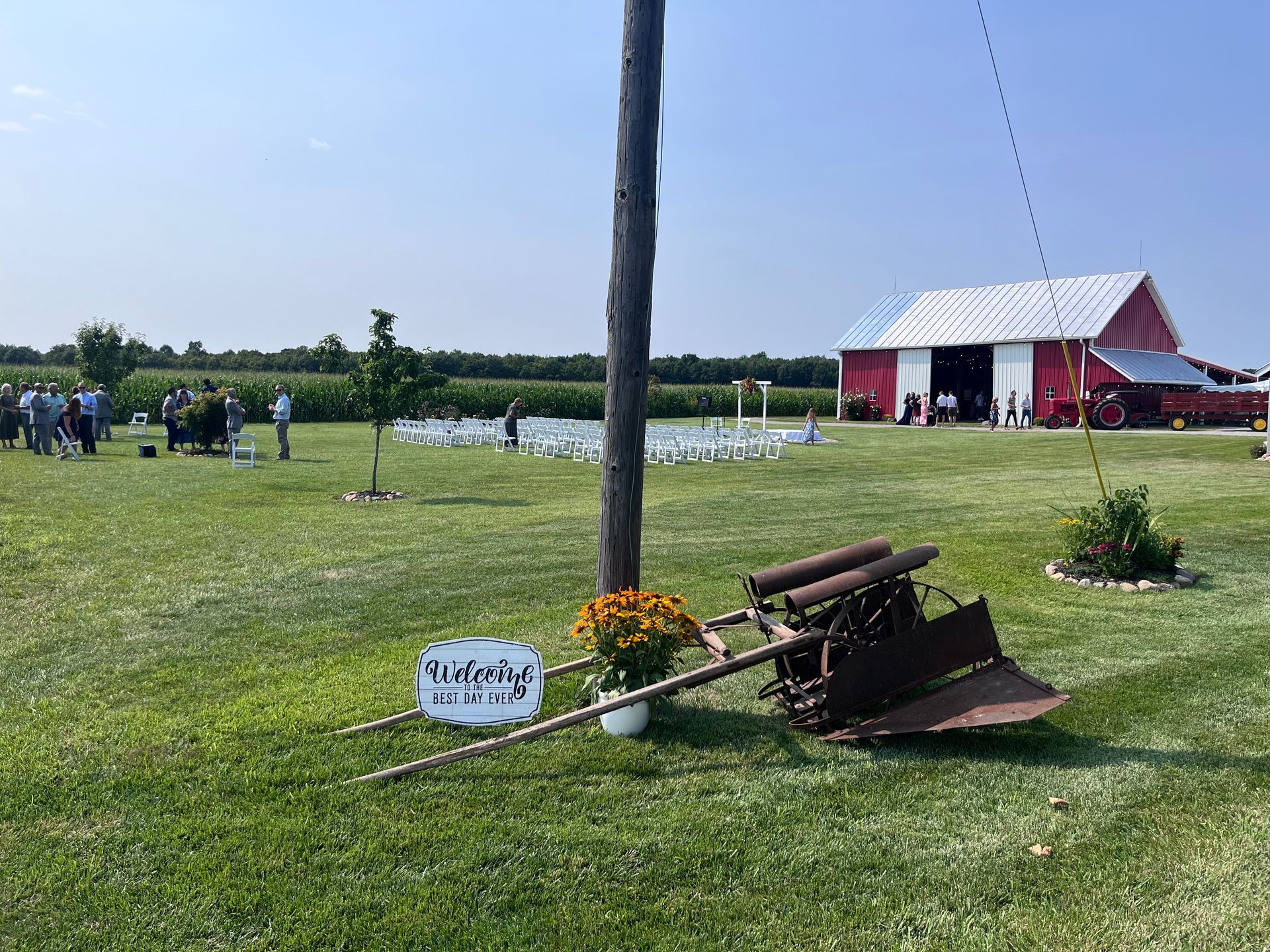 A red barn, farm equipment, and a sign in a grassy field on a sunny day. People gather in the background.